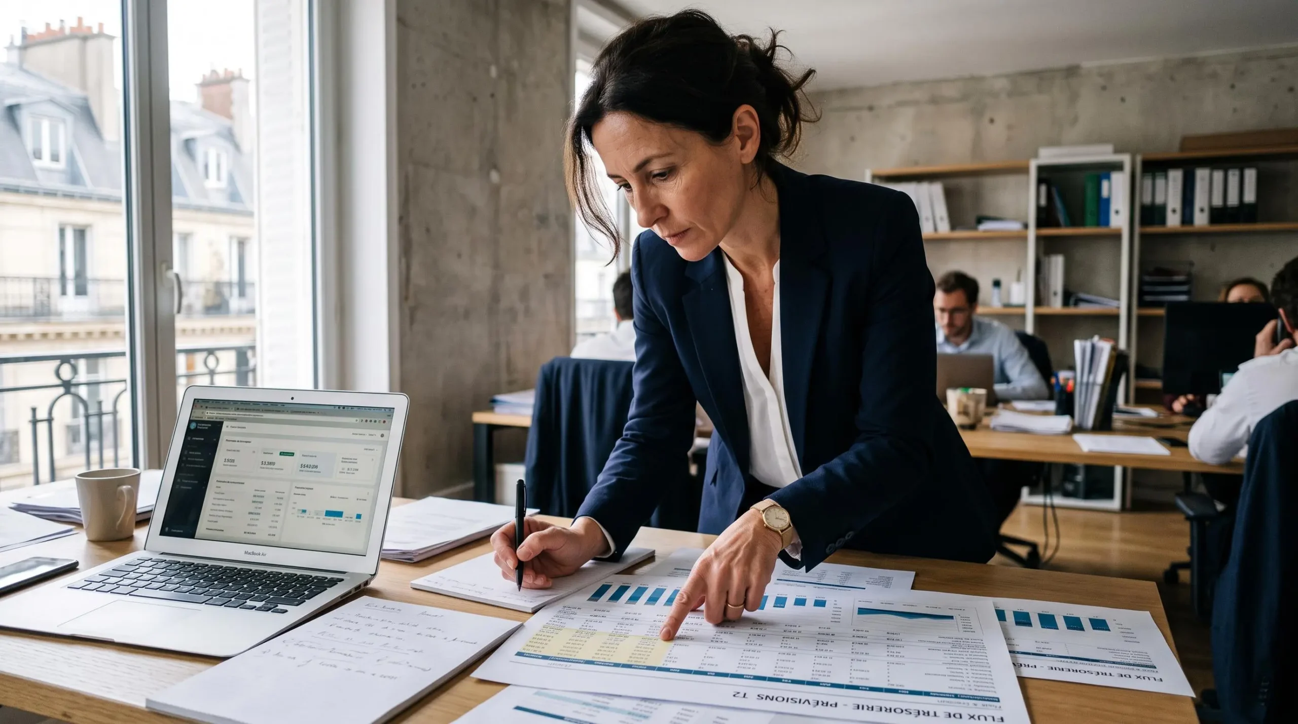 Directeur financier analysant debout un prévisionnel de trésorerie avec plusieurs documents sur son bureau dans un environnement professionnel français.